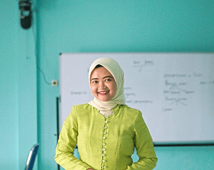 Physics teacher in classroom wearing hijab and green blouse, standing in front of whiteboard, smiling. Physics education concept.