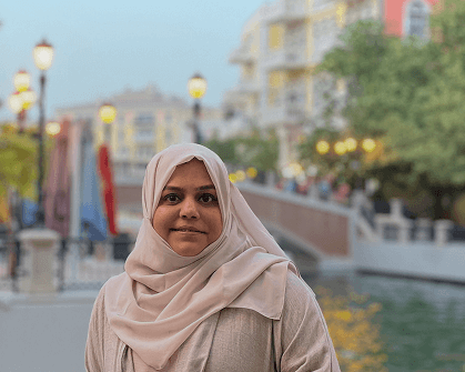 Mathematics tutor portrait: Woman in hijab smiles by canal, European-style buildings in background. Educational resource image.